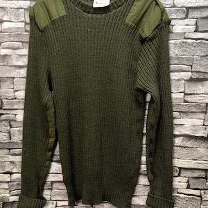 Pode incluir: Um suéter de malha verde com ombros e cotovelos reforçados. O suéter tem gola redonda e é feito de um material grosso e quente.