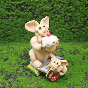 以下が含まれることがあります： 2匹の豚のキャラクターをフィーチャーした気まぐれな置物セット。1匹はさくらんぼのトッピングが付いたカップケーキを持ち、もう1匹は「Diet Way」と書かれた本を噛んでいます。フィギュアは本の上にあり、緑の苔の背景に設定されています。