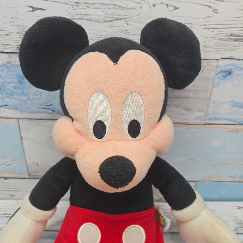 K&ouml;nnte beinhalten: Ein Mickey-Mouse-Pl&uuml;schtier mit schwarzem Kopf, gro&szlig;en runden Ohren, pfirsichfarbenem Gesicht und schwarzer Nase. Die Figur tr&auml;gt ein rotes Unterteil mit zwei wei&szlig;en runden Kn&ouml;pfen sowie wei&szlig;e Handschuhe an den H&auml;nden.