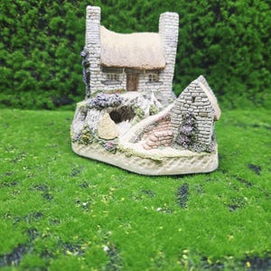 Puede incluir: Una casa de piedra en miniatura con tejado de paja, chimeneas y pequeñas ventanas. La casa está situada sobre una base con un camino y un pequeño edificio. La escena está rodeada de césped verde artificial.