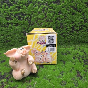 以下が含まれることがあります： 両手を上げたピンク色の豚の置物。黄色い箱には「Piggir」と「Piggin' Toothache」の文字が書かれており、豚のイラストが描かれています。背景は緑色です。