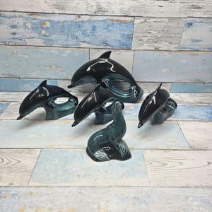 Peut inclure: Un ensemble de cinq figurines en céramique brillante comprenant quatre dauphins noirs dans diverses postures de nage et une otarie dressée. Chaque pièce présente une finition lisse et réfléchissante avec des dégradés de bleu sarcelle et de noir.