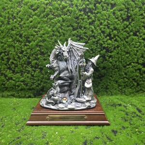 Puede incluir: Figura de fantasía detallada que representa un dragón, un mago y una pequeña criatura. El dragón tiene las alas extendidas y está junto al mago, que sostiene un bastón. La figura está montada sobre una base de madera con el texto "the taskmaster".