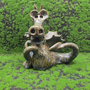 Puede incluir: Escultura de dragón de cerámica con esmalte verde y marrón. El dragón tiene ojos grandes, un hocico ancho y alas detalladas. Posado sobre un lecho de musgo verde, creando una pieza decorativa de fantasía.