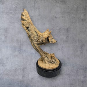 May include: A detailed sculpture of a fox crafted from wood-like material. The fox is perched atop a black, circular base. The sculpture has a natural, textured appearance, with the fox's head and body extending upwards. The base is approximately 3 inches in diameter.