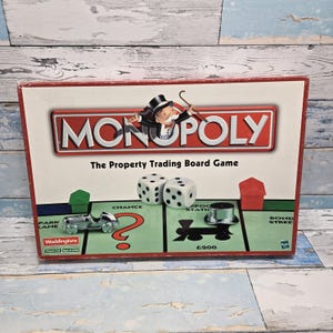 Könnte beinhalten: Monopoly-Brettspielbox mit dem Spieletitel in einem rot-weißen Rahmen. Die Box zeigt Spielfiguren, Würfel und den Monopoly-Mann. Das Spiel wird von Waddingtons hergestellt.