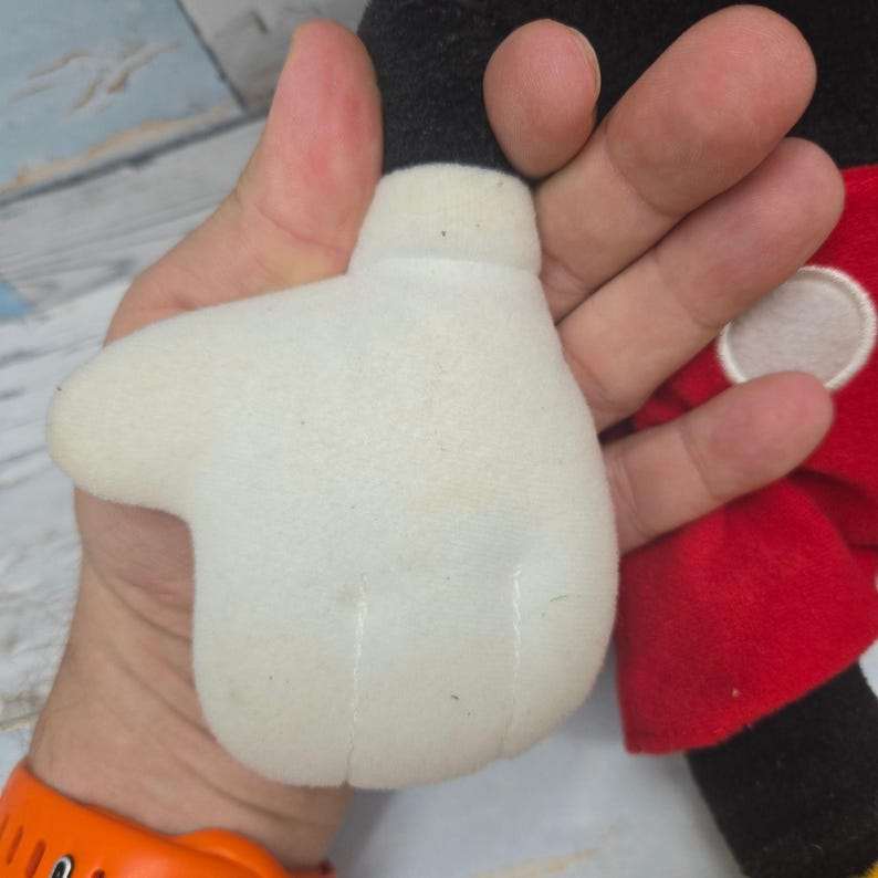 K&ouml;nnte beinhalten: Ein Pl&uuml;schtier mit einer gro&szlig;en, wei&szlig;en, vierfingrigen Cartoon-Handschuhhand aus weichem Stoff. Das Spielzeug hat einen schwarzen Armbereich und einen leuchtend roten Stoffteil mit einem kreisf&ouml;rmigen, wei&szlig;en Knopfdetail an der Seite der Figur.