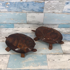 May include: Two intricately carved wooden turtle figurines, both dark brown in colour, are displayed on a light blue and white woodgrain background. The turtles have detailed carvings on their shells and legs.