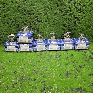 Könnte beinhalten: Kleine silberne Drachenfiguren auf blauen Schachteln mit weißem Text und Barcodes. Die Schachteln sind auf einer grünen, strukturierten Oberfläche angeordnet. Jede Schachtel hat eine andere Produktnummer, wie z. B. 3504, 3535, 3572, 3576, 3574 und 3525.