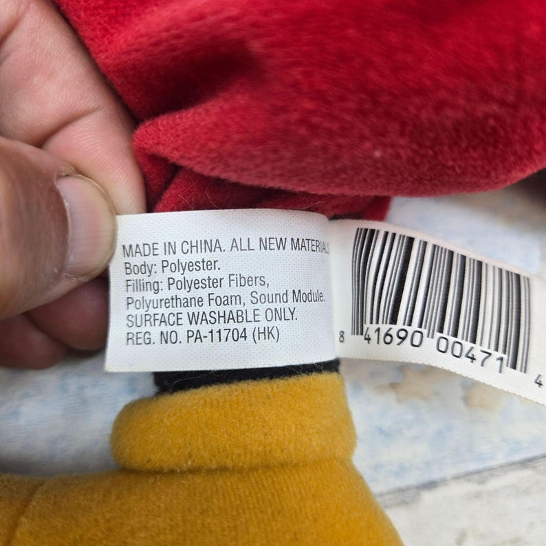 K&ouml;nnte beinhalten: Ein wei&szlig;es Produktetikett aus Stoff mit schwarzem Text, der MADE IN CHINA. ALL NEW MATERIAL Body: Polyester. Filling: Polyester Fibers, Polyurethane Foam, Sound Module. SURFACE WASHABLE ONLY. REG. NO. PA-11704 (HK) neben einem Strichcode lautet.