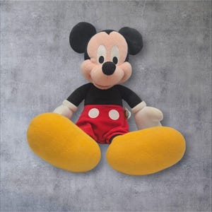 K&ouml;nnte beinhalten: Ein Mickey-Mouse-Pl&uuml;schtier mit schwarzem Kopf und gro&szlig;en runden Ohren, pfirsichfarbenem Gesicht und schwarzer Nase. Die Figur tr&auml;gt ein schwarzes Langarmshirt, eine rote Hose mit zwei wei&szlig;en runden Kn&ouml;pfen, gro&szlig;e gelbe Schuhe und wei&szlig;e Handschuhe.
