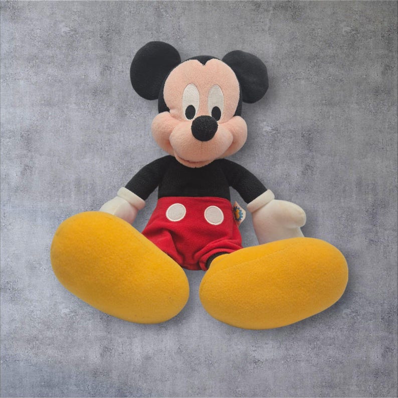 K&ouml;nnte beinhalten: Ein Mickey-Mouse-Pl&uuml;schtier mit schwarzem Kopf und gro&szlig;en runden Ohren, pfirsichfarbenem Gesicht und schwarzer Nase. Die Figur tr&auml;gt ein schwarzes Langarmshirt, eine rote Hose mit zwei wei&szlig;en runden Kn&ouml;pfen, gro&szlig;e gelbe Schuhe und wei&szlig;e Handschuhe.