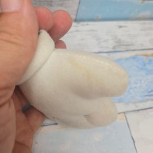 K&ouml;nnte beinhalten: Ein wei&szlig;er Pl&uuml;schhandschuh mit abgerundetem B&uuml;ndchen und drei ausgepr&auml;gten Fingerabschnitten, der eine weiche, samtige Textur und eine leichte gelbliche Verf&auml;rbung auf der Stoffoberfl&auml;che aufweist.