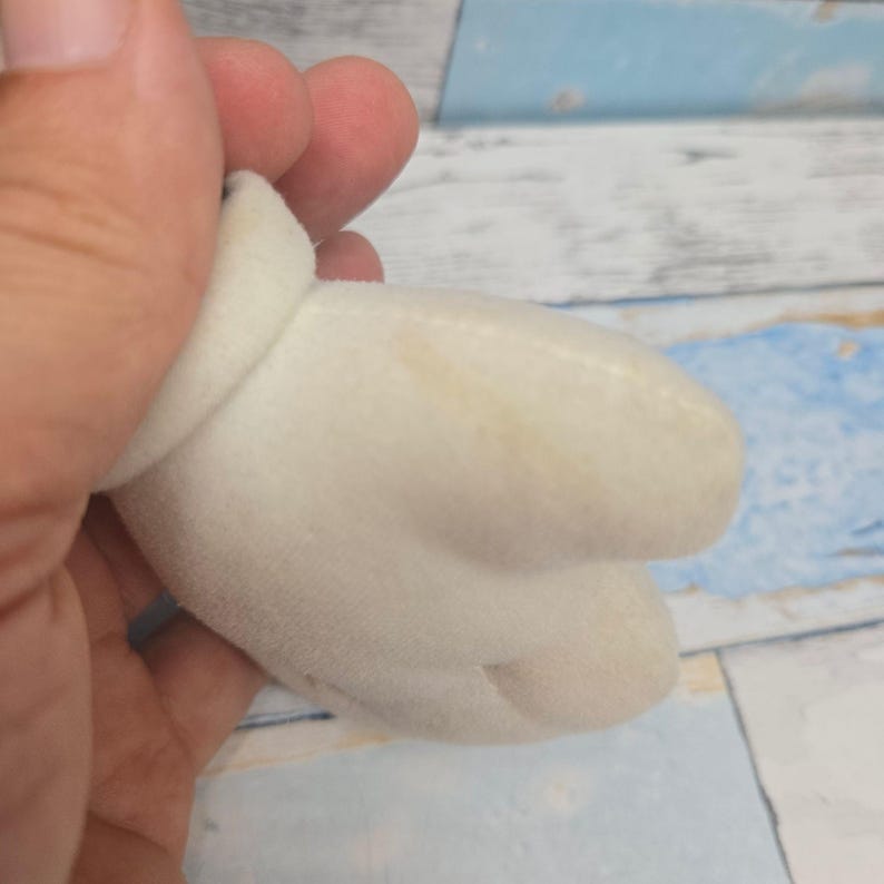 K&ouml;nnte beinhalten: Ein wei&szlig;er Pl&uuml;schhandschuh mit abgerundetem B&uuml;ndchen und drei ausgepr&auml;gten Fingerabschnitten, der eine weiche, samtige Textur und eine leichte gelbliche Verf&auml;rbung auf der Stoffoberfl&auml;che aufweist.