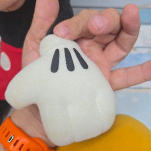 K&ouml;nnte beinhalten: Ein wei&szlig;er Cartoon-Handschuh aus Pl&uuml;sch mit drei schwarzen Ziern&auml;hten auf dem Handr&uuml;cken, befestigt an einem schwarzen Stoff&auml;rmel mit rotem B&uuml;ndchen, das einen wei&szlig;en ovalen Knopf aufweist, und einem gelben, abgerundeten Sockel.