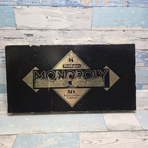 May include: A black box with gold lettering for the 50th Anniversary Edition of the Monopoly board game. The box features the Waddingtons logo and the iconic Monopoly game title.