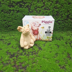 以下が含まれることがあります： 笑顔のベージュ色の豚の置物が、「Piggin」という文字が入った箱の前に置かれています。箱には豚とガチョウのイラストが描かれています。置物は緑色の苔の上に置かれています。