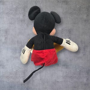 K&ouml;nnte beinhalten: Ein Pl&uuml;schtier einer Zeichentrick-Mausfigur, von hinten betrachtet. Die Figur hat einen schwarzen Kopf und Oberk&ouml;rper, gro&szlig;e runde schwarze Ohren, wei&szlig;e Handschuhe mit drei schwarzen Linien, eine rote Hose und einen langen, d&uuml;nnen schwarzen Schwanz.