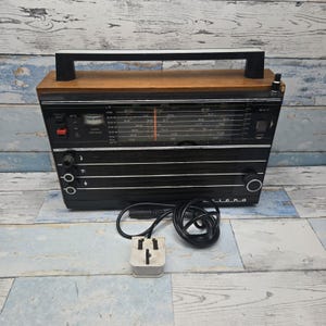 Könnte beinhalten: Ein tragbares Radio mit schwarzer Frontblende, holzfarbener Oberseite und schwarzem Tragegriff. Die Vorderseite verfügt über eine horizontale Frequenzanzeige, eine Sendersuchanzeige, mehrere Drehregler und ein Lautsprechergitter. Ein schwarzes Netzkabel mit Stecker ist angeschlossen.