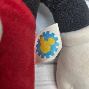 K&ouml;nnte beinhalten: Ein Pl&uuml;schtier mit einem wei&szlig;en Stoffetikett, das ein blaues zahnradf&ouml;rmiges Symbol um eine gelbe Silhouette eines Mauskopfes zeigt. Das Spielzeug hat einen roten Stoffbereich, einen schwarzen flauschigen Arm und eine wei&szlig;e Pl&uuml;schhand.