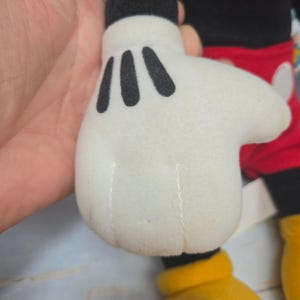 K&ouml;nnte beinhalten: Eine Pl&uuml;schtierhand aus wei&szlig;em Stoff mit drei schwarzen Ziern&auml;hten auf dem Handr&uuml;cken, befestigt an einem schwarzen Arm. Die Figur tr&auml;gt eine rote Hose mit wei&szlig;en ovalen Kn&ouml;pfen und hat gelbe Stofff&uuml;&szlig;e, die am unteren Rand sichtbar sind.