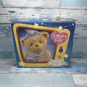 Puede incluir: Una caja azul con una imagen de un oso de peluche con un vestido rosa y un collar de perlas. La caja tiene una etiqueta en forma de corazón que dice "A Bear's Life". La caja es del Club The Cherished Teddies.