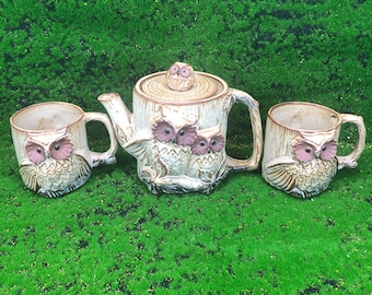 May include: A ceramic owl-themed tea set featuring a teapot and two mugs. The teapot has a tree trunk design with three owl figures and a small owl on the lid. The mugs and teapot are beige with pink and brown accents.