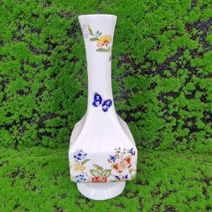 May include: A white ceramic vase with a square base and a tall, slender neck. The vase is decorated with a floral pattern in shades of blue, yellow, red, and green, and features a butterfly design.