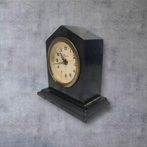 Peut inclure: Une horloge de cheminée vintage en bois noir avec un cadran crème et une lunette dorée. L'horloge présente des aiguilles noires et des chiffres arabes. L'horloge est rectangulaire avec un sommet en pointe et repose sur une petite base.