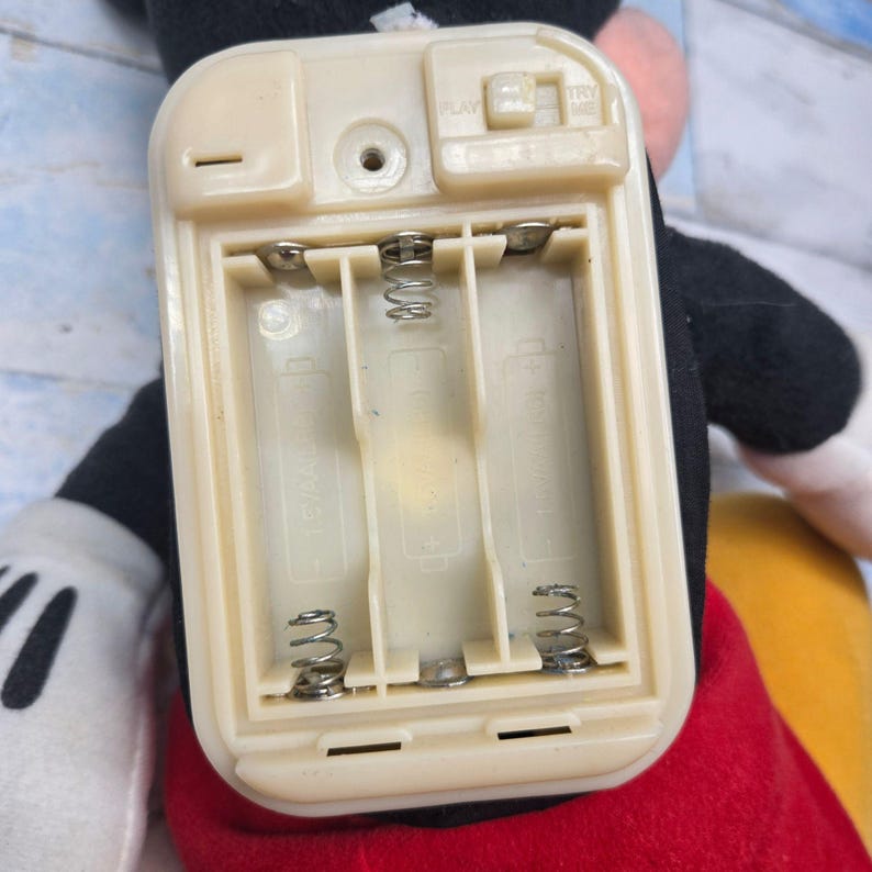 K&ouml;nnte beinhalten: Ein beigefarbenes Kunststoff-Batteriefach f&uuml;r ein Spielzeug mit drei F&auml;chern f&uuml;r AA-Batterien und Metallfedern an der Basis. Der obere Teil enth&auml;lt einen kleinen Schiebeschalter mit der Aufschrift PLAY und TRY ME sowie ein zentrales Schraubloch.