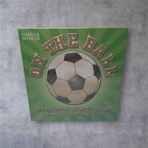 Puede incluir: Una caja de juego de temática futbolística verde y blanca con el texto "ON THE BALL" y un gráfico de balón de fútbol. La caja también presenta el texto "DO YOU HAVE WHAT IT TAKES TO WIN THE WORLD CUP?" y el logotipo de Marks & Spencer.