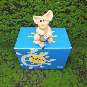 以下が含まれることがあります： 「Piggin' Lite!」の文字と黄色のアクセントが入った青い箱の上に、ベージュ色の豚の置物が座っています。豚は小さな缶を持ち、小さなブーツを履いています。箱は緑色の苔の上にあります。