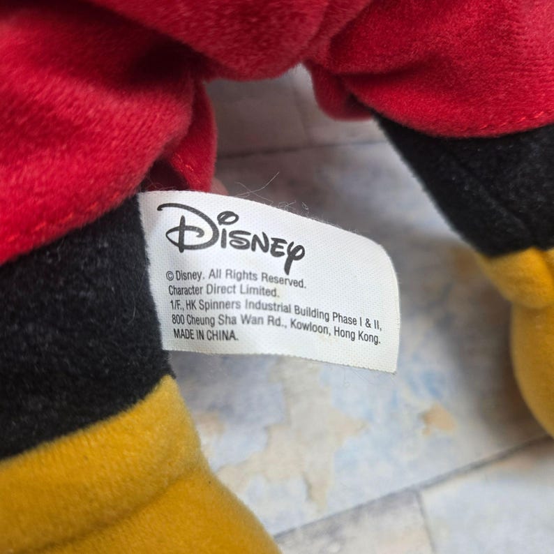 K&ouml;nnte beinhalten: Ein Pl&uuml;schtier mit Abschnitten aus rotem, schwarzem und gelbem Stoff. Ein wei&szlig;es Stoffetikett ist angebracht, das das Disney-Logo und den Text zeigt: Disney. All Rights Reserved. Character Direct Limited. 1/F., HK Spinners Industrial Building Phase I & II, 800 Cheung Sha Wan Rd., Kowloon, Hong Kong. MADE IN CHINA.