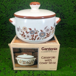 Op de afbeelding: Een witte keramische ovenschaal met een bruin deksel en een bloemenpatroon. De schaal is gelabeld "Gardenia Ceramicware Co-ordinates for the kitchen Casserole with Cover 58 oz. Made in Japan."