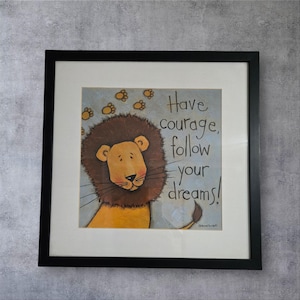 Könnte beinhalten: Gerahmter Kunstdruck mit einer Cartoon-Löwen-Illustration. Der Löwe hat eine braune Mähne und ein freundliches Gesicht. Der Druck enthält den Text "Have courage, follow your dreams!" in handschriftlichem Stil, vor einem hellblauen Hintergrund mit Pfotenabdrücken.