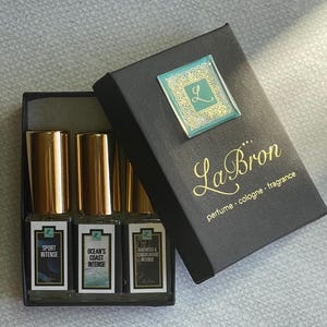 May include: A black box with a gold logo that says "La Bron perfume - cologne - fragrance". The box contains three small glass bottles with gold caps. The bottles are labeled "Sport Intense", "Ocean's Coast Intense", and "Oakmoss & Sandalwood Intense".