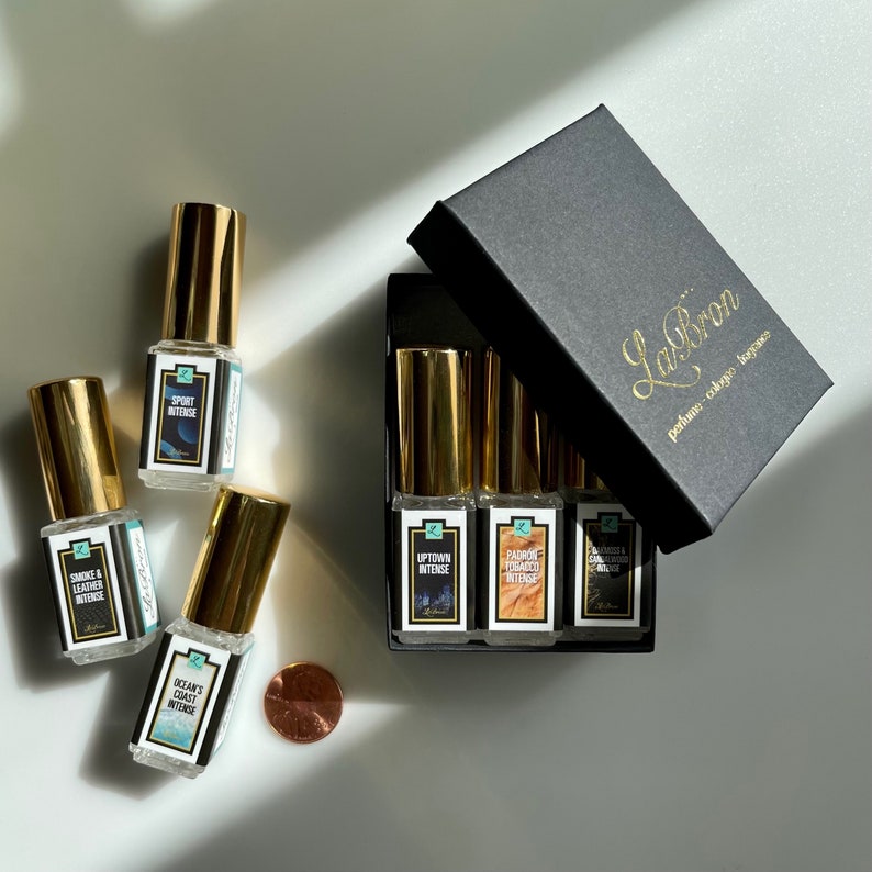 May include: A set of six small glass bottles with gold caps and labels. The bottles are arranged in a black box with the text "LaBron perfume-cologne-bagpace" printed on the top. The labels on the bottles have different names and descriptions of the scents, including "Smoke & Leather Intense", "Sport Intense", "Ocean's Coast Intense", "Uptown Intense", "Padron Tobacco Intense", and "Vanilla & Coconut Intense".