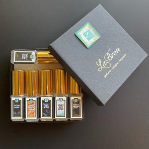 May include: A black box with gold lettering that says "La Bron perfume - cologne - fragrance". The box contains eight small glass bottles with gold caps and labels that say "Intense" and different scents.