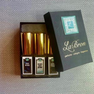 May include: A set of three small gold bottles of perfume in a black box. The bottles are labeled "Sport Intense", "Ocean's Coast Intense", and "Dakmos Sandalwood Intense". The box has the text "La Bron perfume - cologne - fragrance" on it.