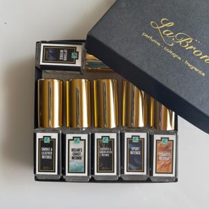 May include: A set of six small glass bottles with gold caps and labels. The labels have different scents, including "Smoke & Leather Intense", "Ocean's Coast Intense", "Darkmoss & Sandalwood Intense", "Sport Intense", and "Tobacco Intense".