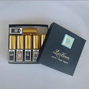 May include: A black box with gold lettering that reads "La Bron perfume-cologne-fragrance". The box contains eight small glass bottles with gold caps and labels that include the names "Uptown Intense", "Padron Tobacco Intense", "Sport Intense", and "Ocean's Coast Intense".