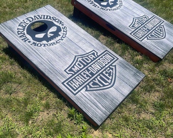 May include: Two wooden cornhole boards with a grey wood grain finish. Each board features Harley-Davidson logos and skull graphics in black. The boards are set on green grass, ready for a game.