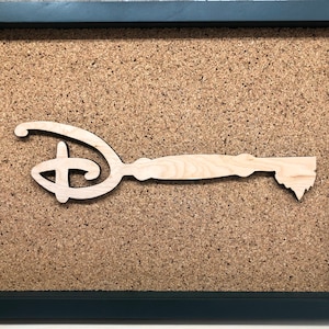 May include: A wooden key with a Disney logo cut out in the shape of the keyhole. The key is displayed in a dark gray frame on a corkboard background.