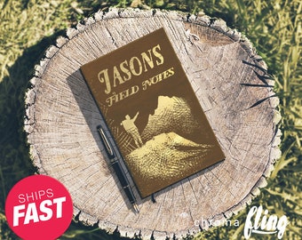 May include: A brown leather notebook with the text "Jasons Field Notes" on the cover. The notebook is sitting on a tree stump with a pen next to it.