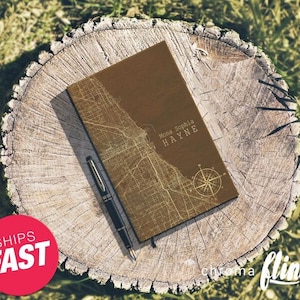 May include: A brown leather journal with a gold map design and the name "Mona Sophia HAYNE" printed on the cover. The journal is sitting on a tree stump with a pen next to it.