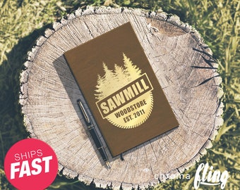 May include: A brown leather notebook with a gold logo that says "Sawmill Woodstore Est. 2011". The logo features a silhouette of pine trees.