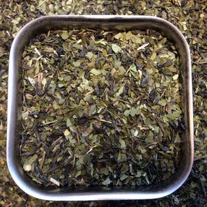 May include: A close-up of a metal container filled with loose leaf green tea. The tea is a blend of green and black tea leaves, with some herbs and flowers mixed in.