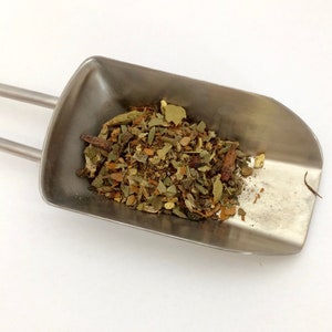 May include: A metal scoop filled with a blend of dried herbs and spices, including green leaves, brown pieces, and orange bits.