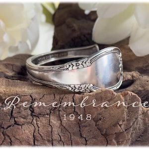 May include: A silver spoon bracelet with floral and ornate detailing. The bracelet is engraved with the word "Remembrance" and the year "1948". The bracelet is displayed on a piece of wood.