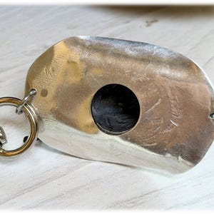 May include: A silver-toned metal dog tag with a smooth, reflective surface and a central circular cutout. The tag features a small hole on the right side and is attached to a circular metal split ring on the left side, creating a keychain component.